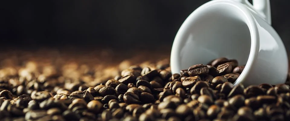 A cup of coffee tipped over in the middle of many coffee beans.