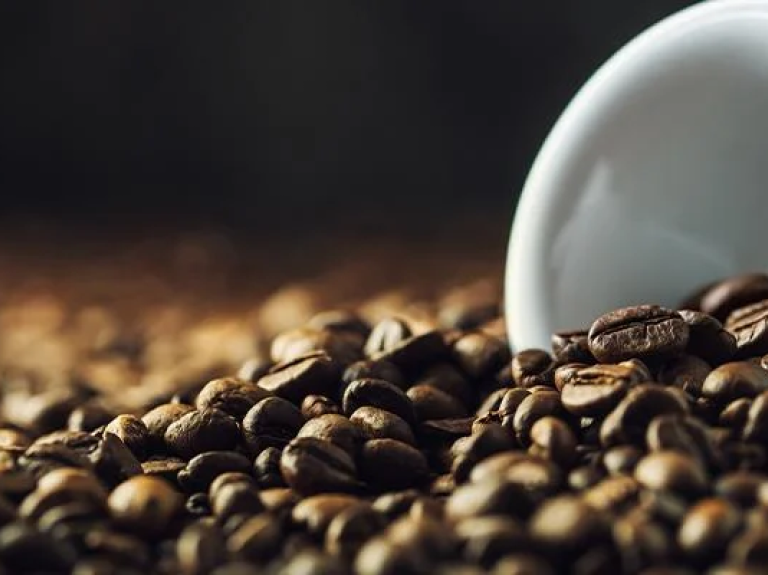 A cup of coffee tipped over in the middle of many coffee beans.