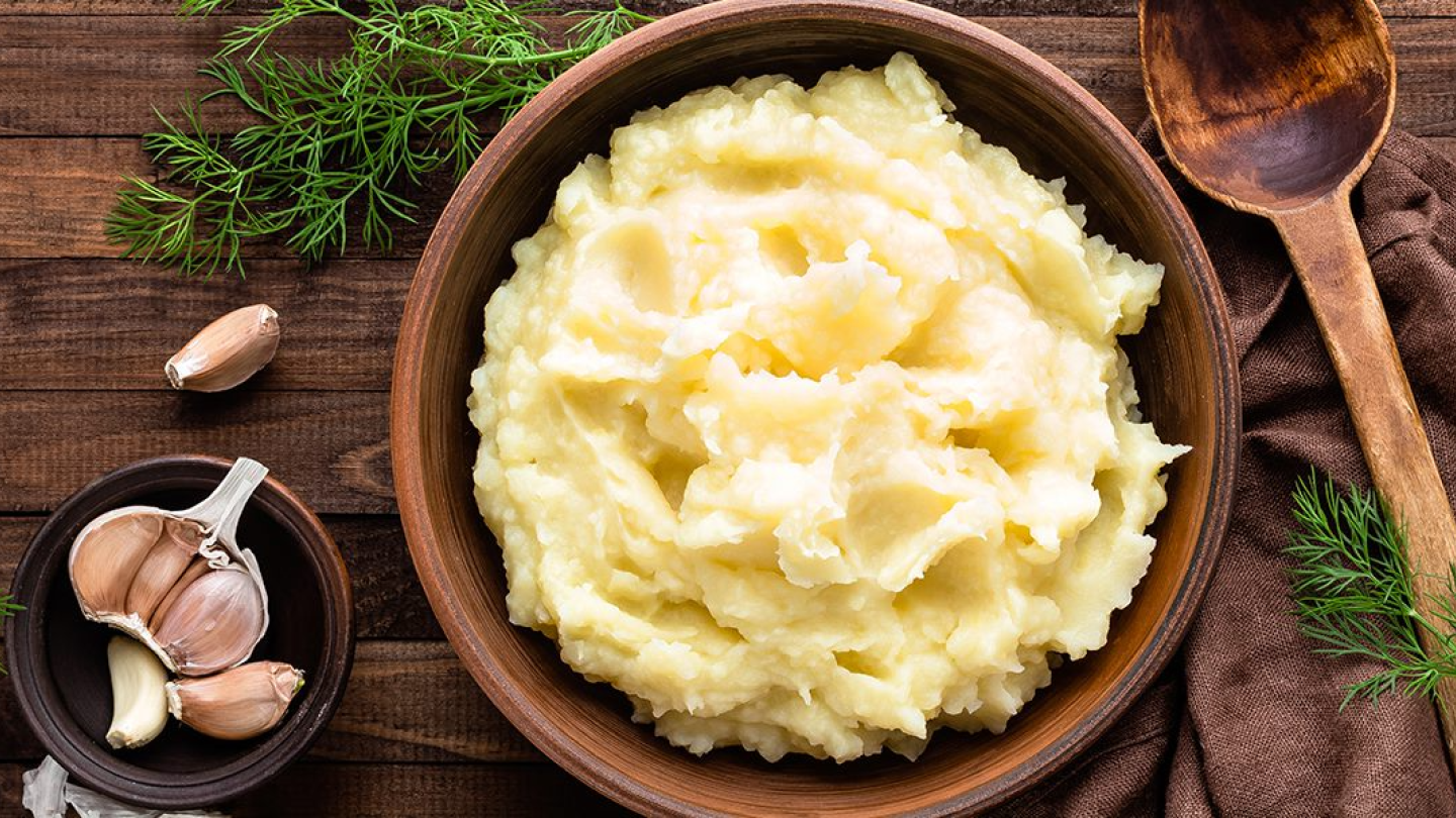 A plate of mashed potatoes on a table with garlic and rosemary on the side.