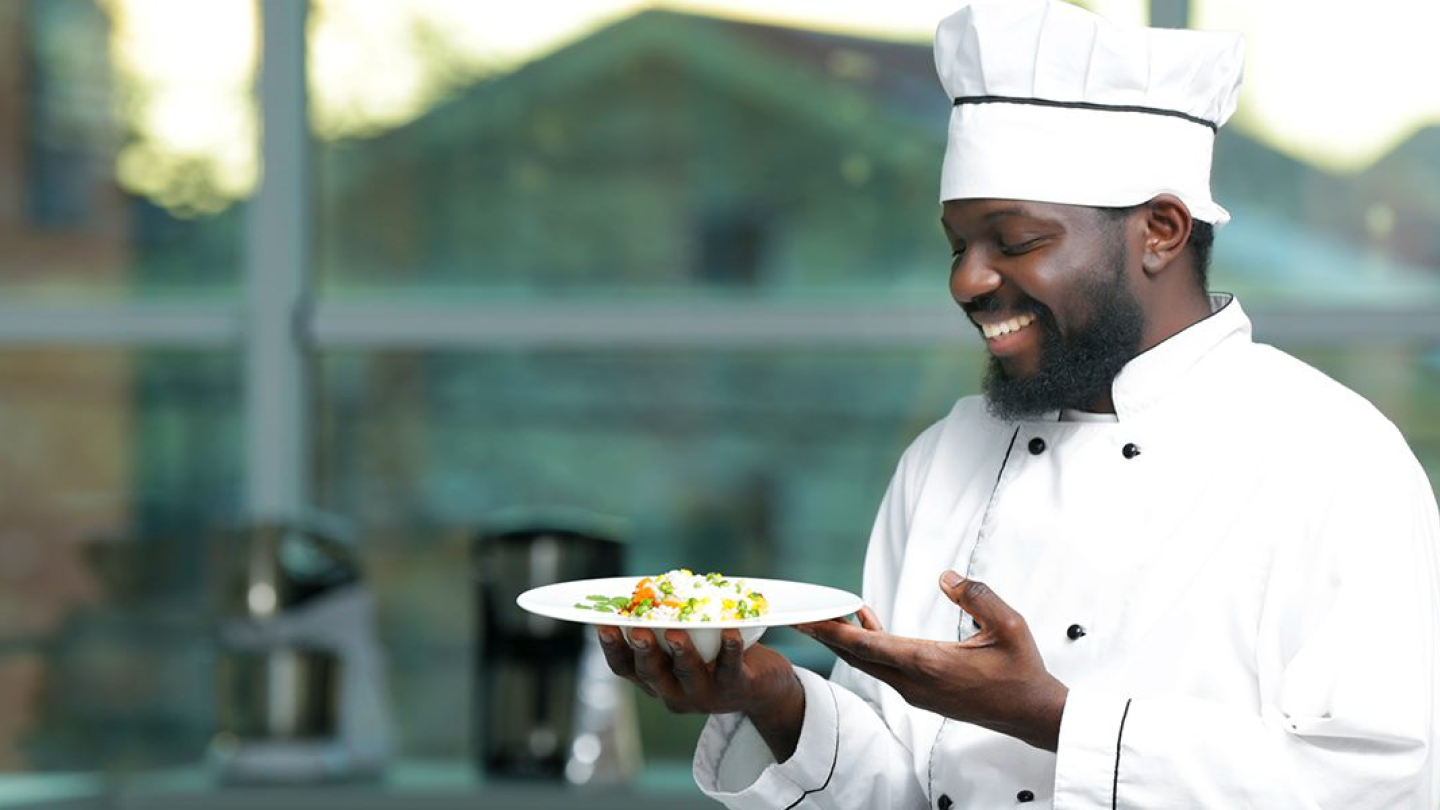 A male chef smiling at a plate of food he is holding in his hands.